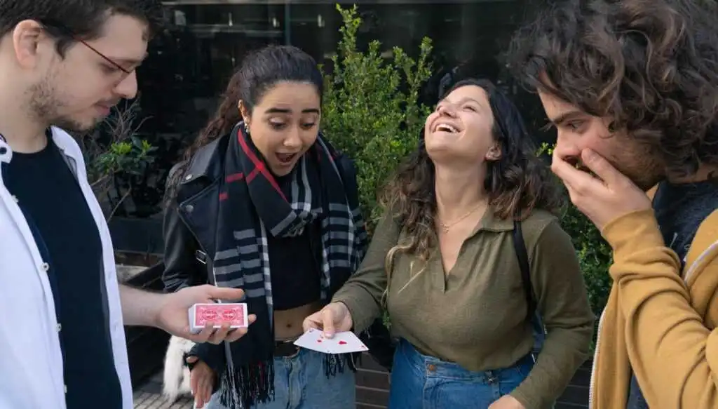 Three young people show fascination and laughter, one of them has two cards in her hand and in front of them Maximiliano Giaconia holds a deck of cards.