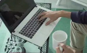 A man indoors in front of his laptop, one hand on the keyboard and holding a cup with the other.